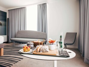 A modern hotel room with a grey sofa, a dining table with food and drinks, a chair, and a large window with curtains. - Novotel Paris Coeur D'orly (Orly)