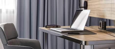 Ein modernes Zimmer mit Schreibtisch, Laptop, Stuhl und Vorhängen.