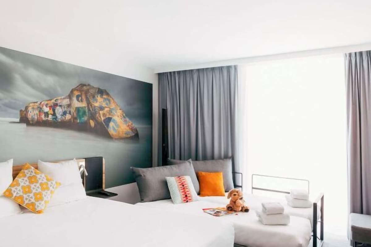 Habitación de hotel con cama, escritorio, silla, un cuadro en la pared y ventana con cortinas.
