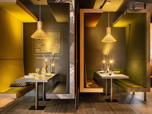 A modern interior with two dining areas, each featuring a table, bench, pendant lighting, and a wall with a cityscape illustration. - Novotel Paris Coeur D'orly (Orly)