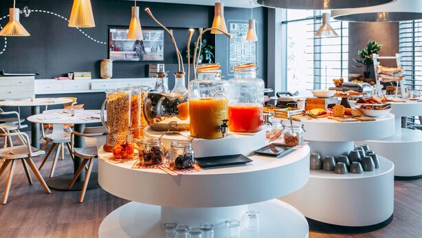 A modern breakfast buffet with various food items and beverages, contemporary furniture, and large pendant lights. - Novotel Paris Coeur D'orly (Orly)