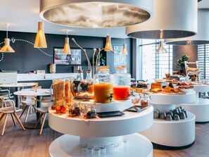 A modern breakfast buffet with various food items and beverages, contemporary furniture, and large pendant lights. - Novotel Paris Coeur D'orly (Orly)