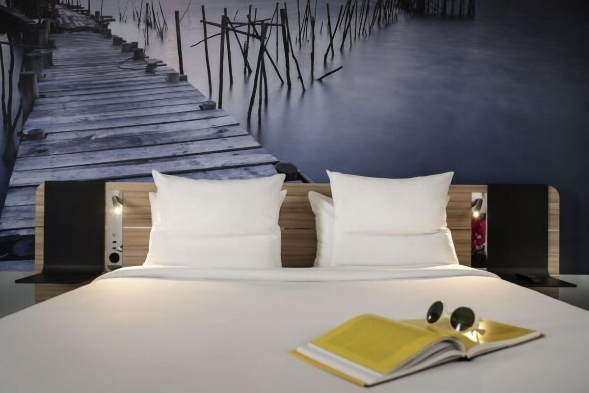 Una cama con ropa blanca, dos almohadas, un libro y un par de gafas sobre la cama.