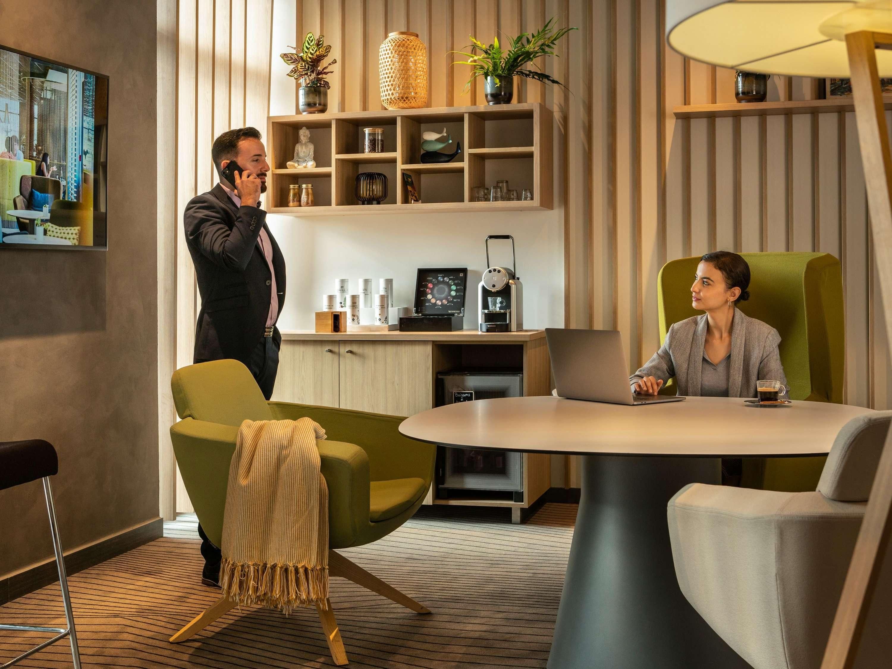A modern hotel room with a round table, two chairs, a coffee machine, and a shelf with decorative items.
