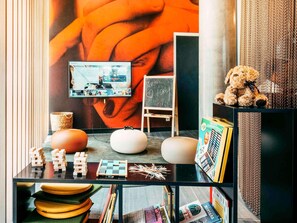 Una habitación con un televisor grande, un caballete pequeño, un osito de peluche y una estantería con libros y juegos de mesa.