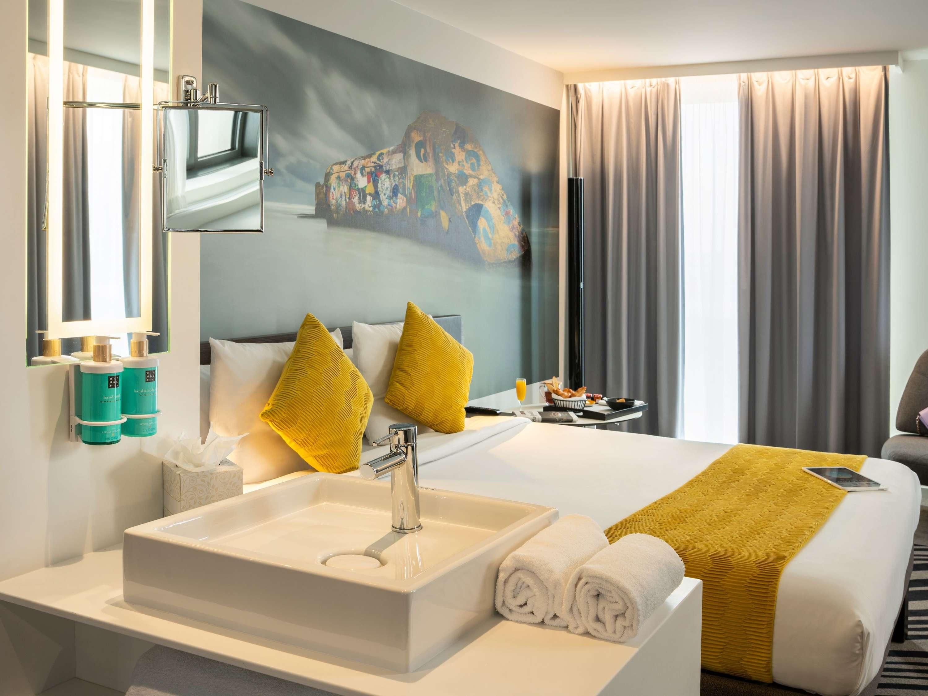 Habitación de hotel con una cama grande, un lavamanos blanco, un espejo y un cuadro en la pared.
