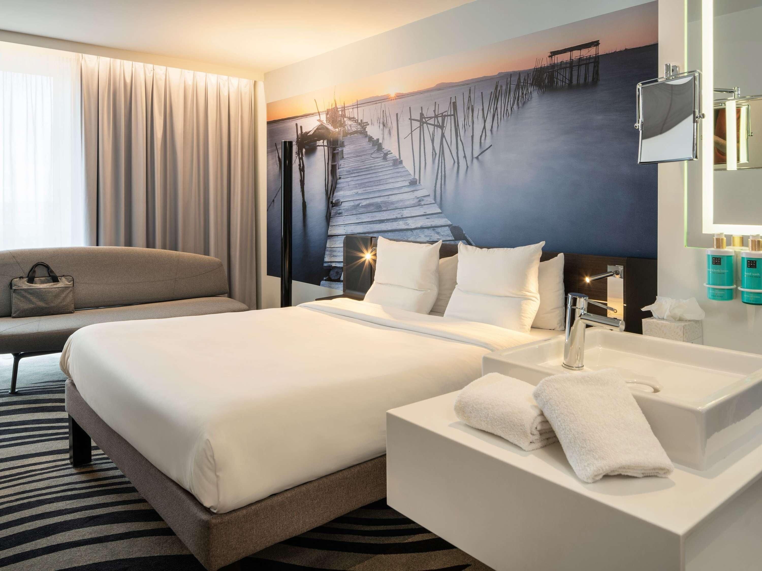 A hotel room with a bed, a sofa, a bathroom with a sink and mirror, and a large window with curtains.