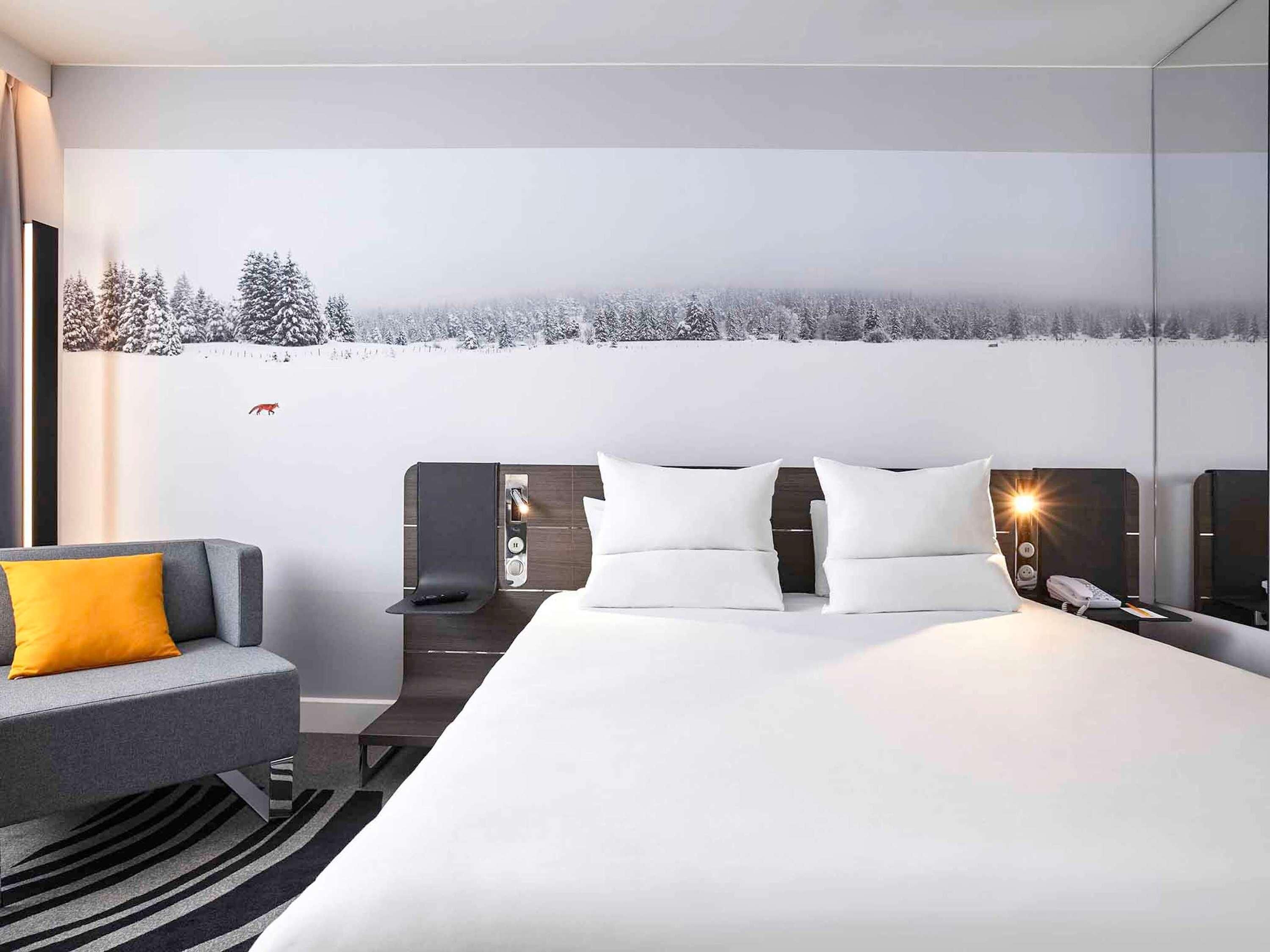 Een moderne hotelkamer met een groot bed, een grijze bank met een geel kussen, een nachtkastje met een lamp en een muurschildering van een besneeuwd landschap.