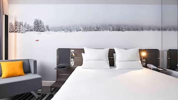 Een moderne hotelkamer met een groot bed, een grijze bank met een geel kussen, een nachtkastje met een lamp en een muurschildering van een besneeuwd landschap.