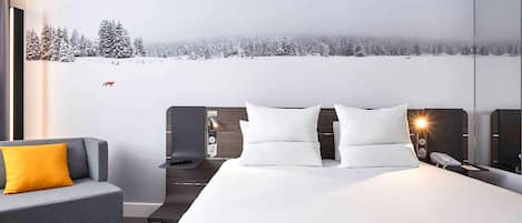 A modern hotel room with a large bed, a gray sofa with a yellow pillow, a nightstand with a lamp, and a wall mural of a snowy landscape.