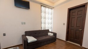 A living room with a dark brown sofa, a patterned rug, a wooden floor, a large window with lace curtains, and a dark wooden door. - Hotel Rivas  (Tequila)