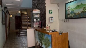 A hotel reception area with a wooden reception desk, a staircase, a wall-mounted emergency exit sign, and a painting of a landscape. - Hotel Rivas  (Tequila)