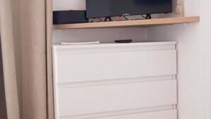 A room with a flat-screen TV on a wooden shelf, a white drawer unit below, and a beige curtain on the left. - Studio number 15 in Pantin (Pantin)