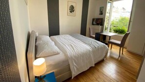 Ein Schlafzimmer mit Bett, Nachttisch, Lampe und einem Fenster mit Blick auf Grünflächen und Gebäude.