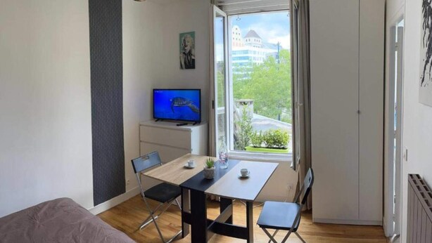 Ein kleiner, moderner Raum mit einem Holztisch, zwei Stühlen, einem Fernseher auf einem weißen Schrank und einem großen Fenster mit Blick auf Gebäude und Grünflächen.