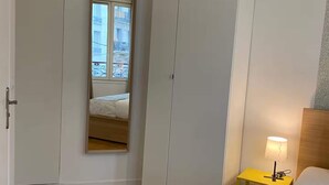 A bedroom with a wooden floor, a bed with a white headboard, a nightstand with a lamp, a wardrobe, and a mirror reflecting a window view. - Studio number 6 in Pantin (Pantin)