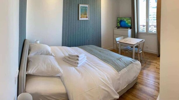 Een netjes opgemaakt bed met witte lakens en een opgevouwen handdoek, een klein bureau met een stoel, en een raam met uitzicht op gebouwen.