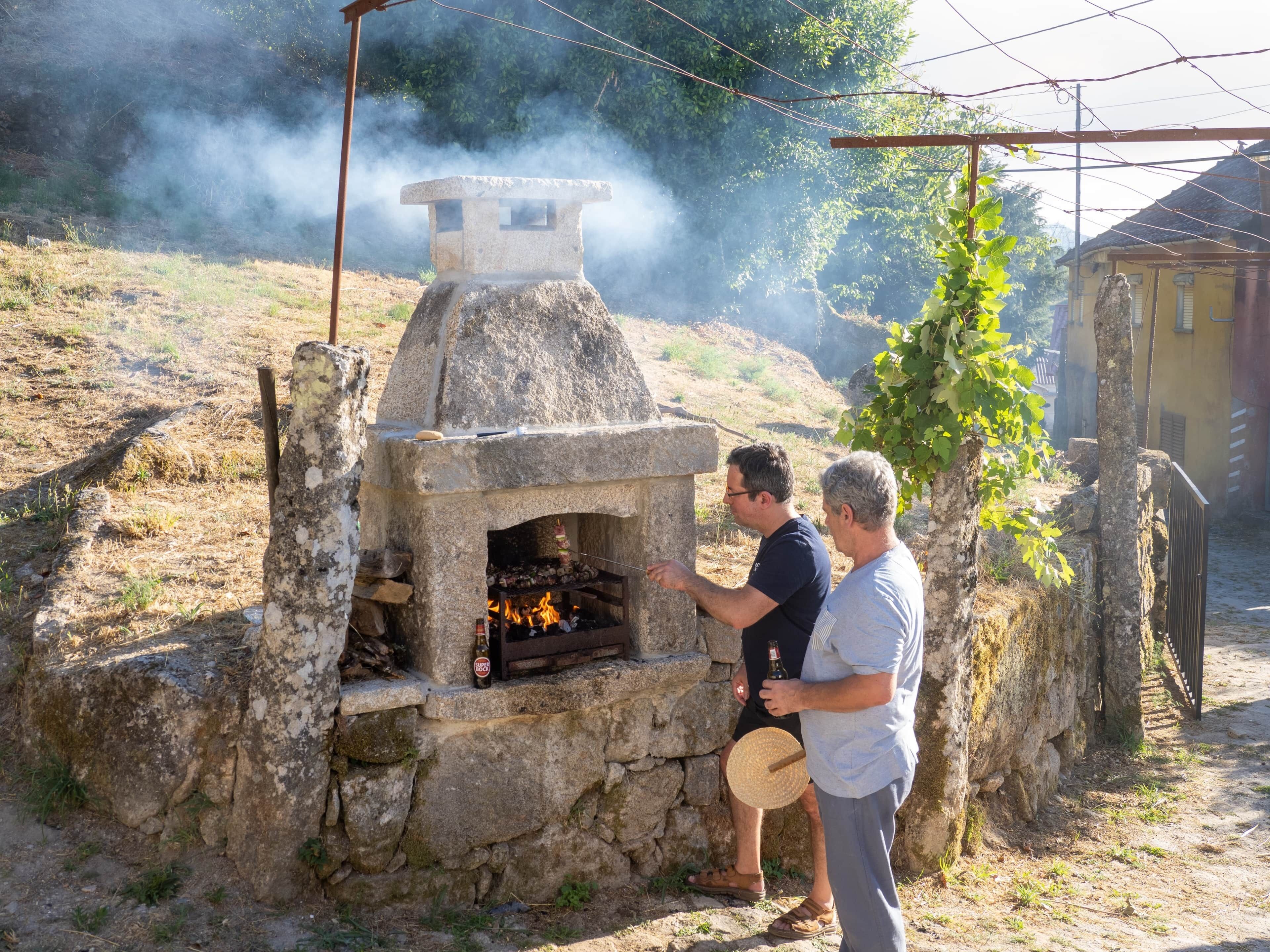 A stone-built outdoor oven with smoke, surrounded by a rustic setting with a tree and a building in the background.