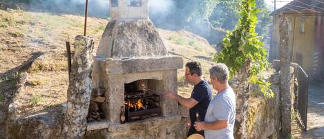 A stone-built outdoor oven with smoke, surrounded by a rustic setting with a tree and a building in the background.