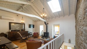 Un vaste salon doté d’un haut plafond, de poutres en bois, d’un mur en pierre, d’une cheminée, d’un coin repas et d’un escalier.