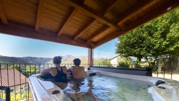 Un bain à remous où se trouvent deux personnes, entouré d’une terrasse en bois et offrant une vue sur les montagnes.
