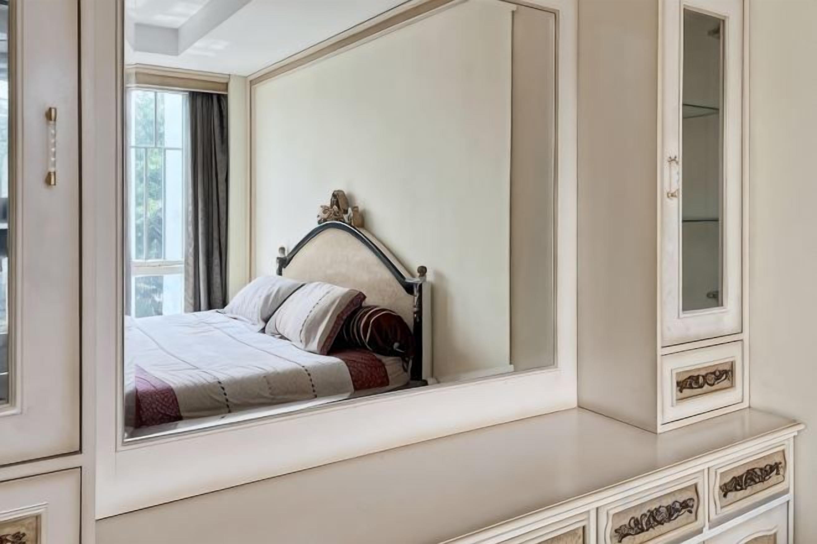 A bedroom with a large mirror reflecting a bed, a dresser with drawers, and a window with curtains.