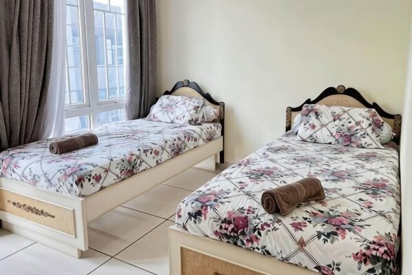Two single beds with floral bedding and wooden headboards in a room with a large window and curtains.