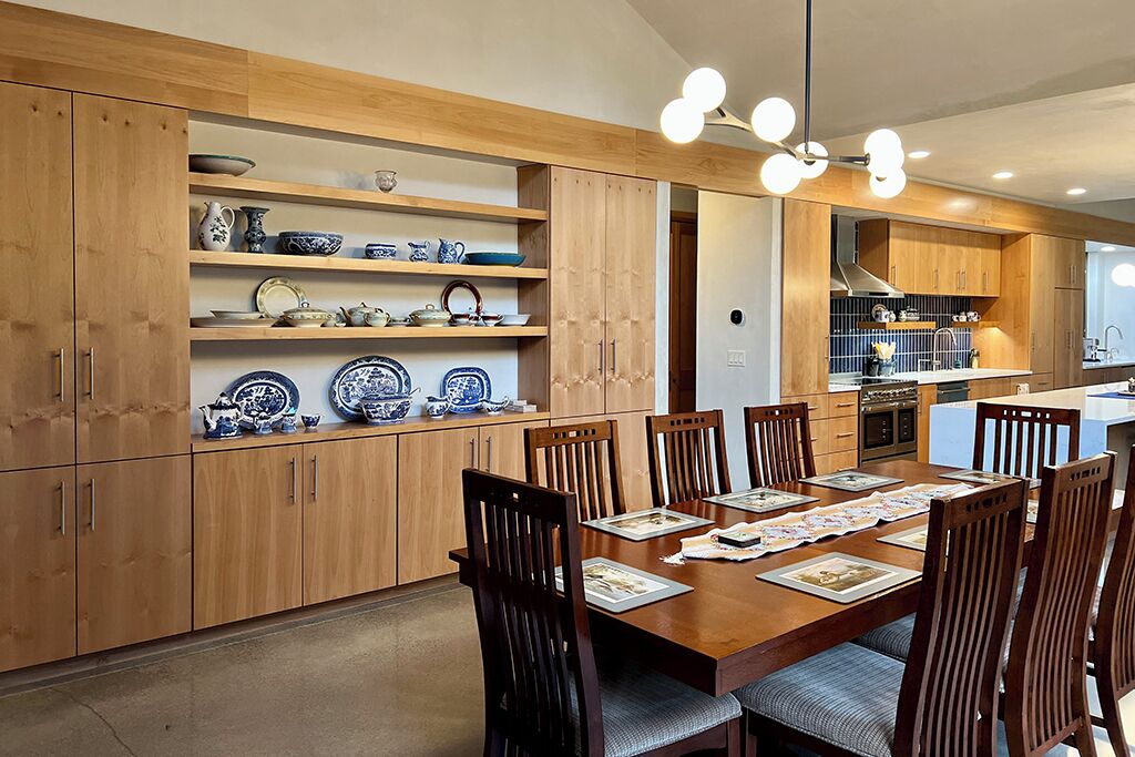 A modern kitchen with wooden cabinets, a dining table set for six, and a built-in shelving unit displaying blue and white dishes.