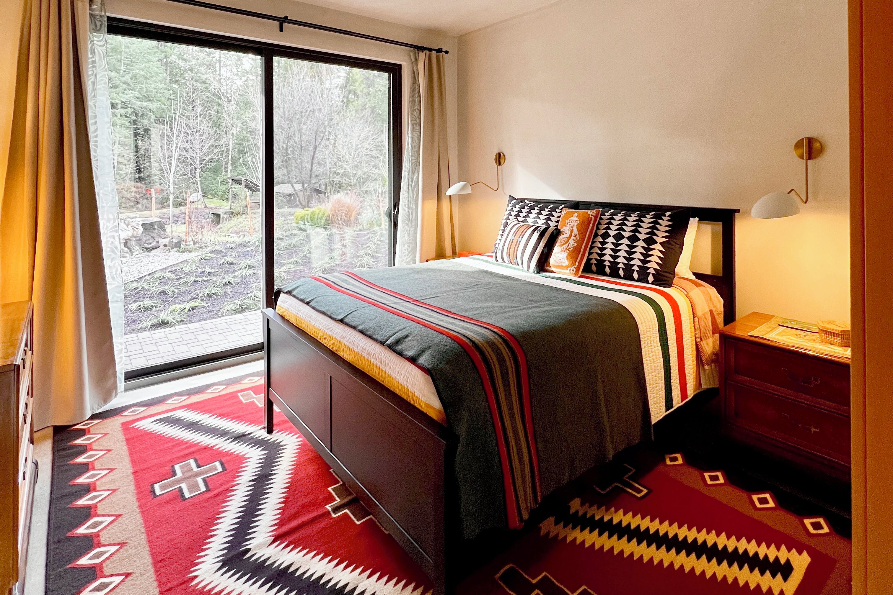 A bedroom with a large window, a bed with patterned bedding, a nightstand, and a wooden dresser.