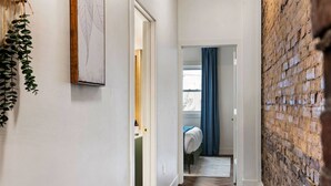 A narrow hallway with a wooden floor, a brick wall on the right, and a bed with a blue curtain in the background. - 2615 Jane Down - Southside (Pittsburgh)