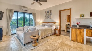 A spacious bedroom with a large bed, a ceiling fan, a view of lush greenery outside, and a modern kitchenette.