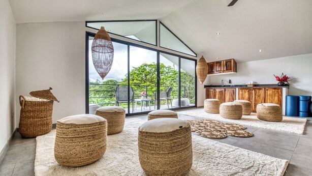 A spacious living area with large windows, woven ottomans, and a modern kitchen.