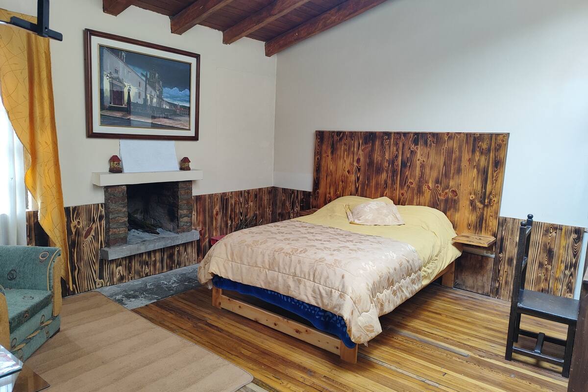 Een slaapkamer met een houten hoofdbord, een bed met een beige en blauwe spreideken, een kleine open haard en een ingelijst schilderij aan de muur.