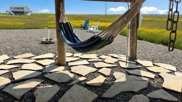 A hammock under a wooden structure, a stone-tiled patio, and a distant view of a house in a grassy field.