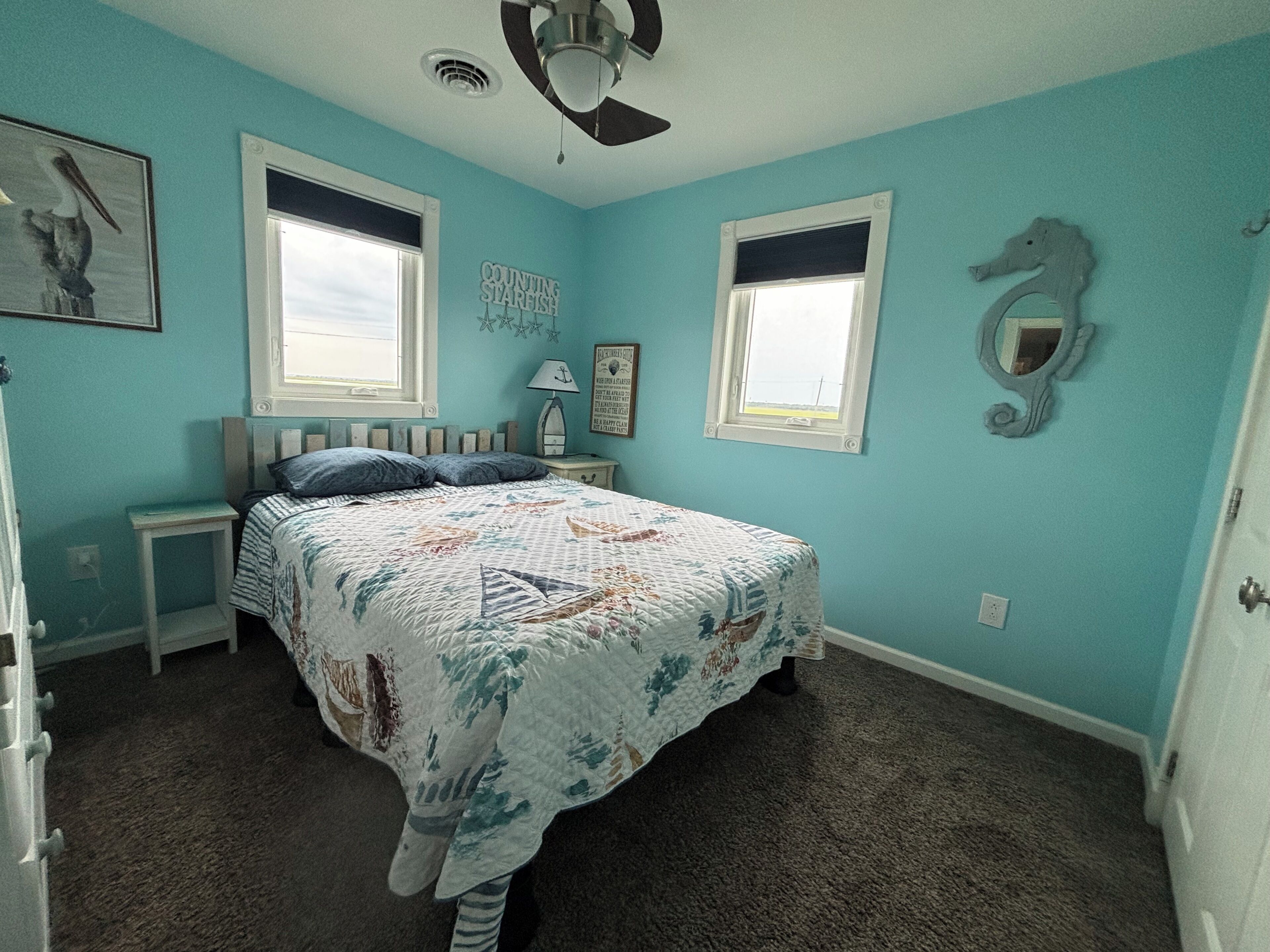 A bedroom with a bed, two nightstands, a ceiling fan, two windows, and a seahorse decoration.