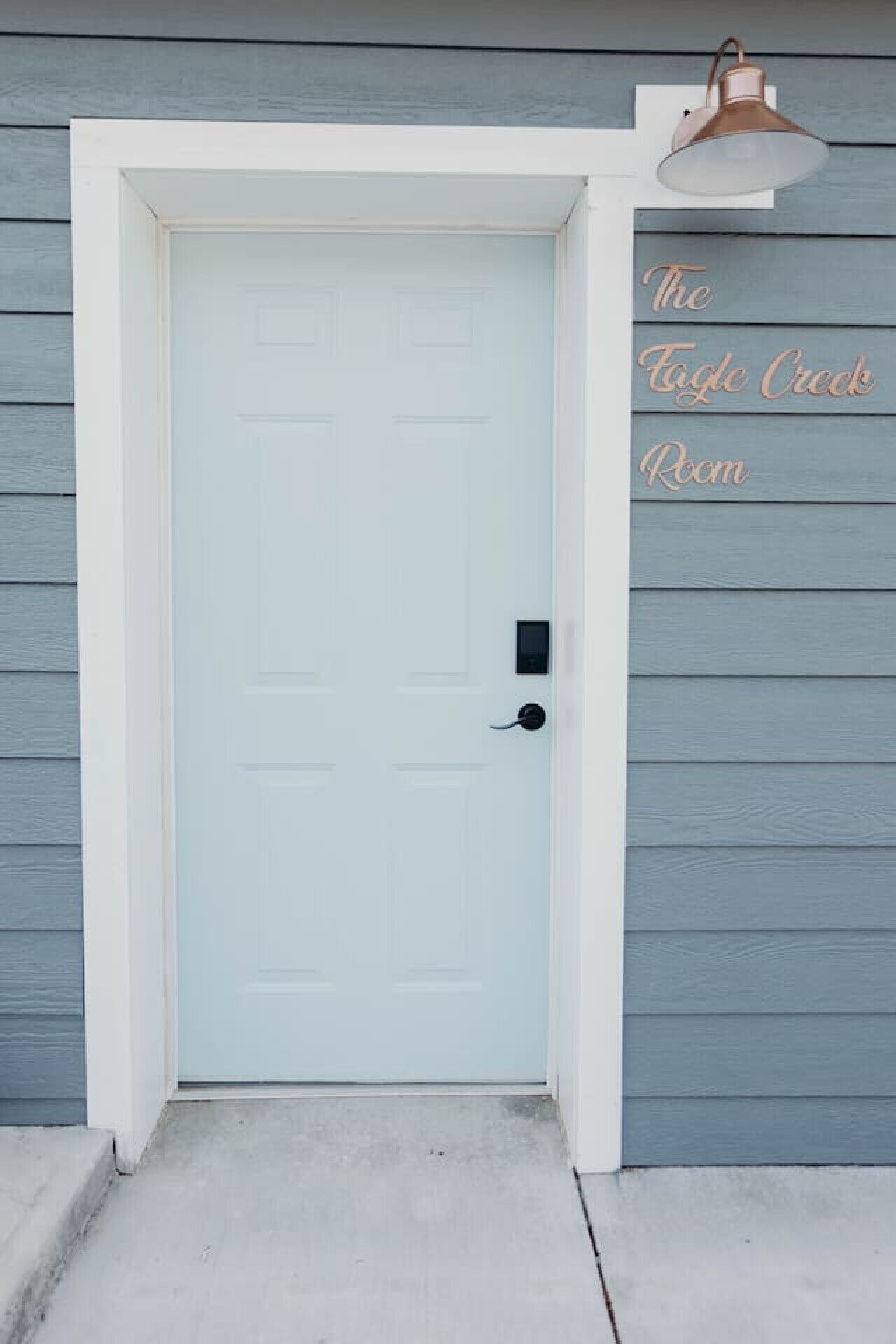 A light blue door with a black handle and a small electronic lock. The door is set in a white frame, and there is a light fixture above it. The wall is painted in a light blue color, and the text "The Eagle Creek Room" is visible on the wall to the right of the door.