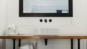 Un baño con pared de azulejos blancos, tocador de madera con espejo y lavamanos.