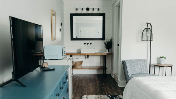Un dormitorio con televisión, cómoda, mesita de noche, silla y un espejo grande.