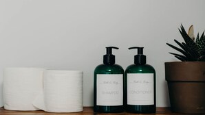 Dos botellas verdes, una con la etiqueta 'Bath & Body SHAMPOO' y otra con 'Bath & Body CONDITIONER', sobre una superficie de madera, junto a una toalla blanca y una planta en maceta.