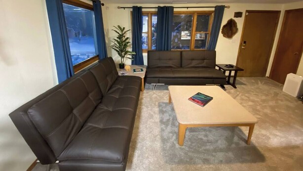 Una sala de estar con un sofá de cuero marrón, una mesa de centro color beige y un sofá marrón oscuro. Se ve una ventana con cortinas azules, una planta en maceta y una puerta al fondo.