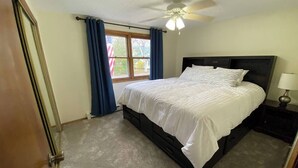 Un dormitorio con cama, ventilador de techo, ventana con cortinas y mesita de noche con lámpara.