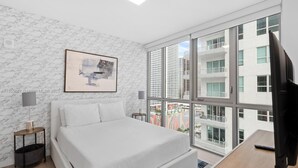 Et moderne soveværelse med en stor seng, et indrammet abstrakt billede, et fjernsyn og udsigt over byens skyline gennem gulv til loft vinduer.