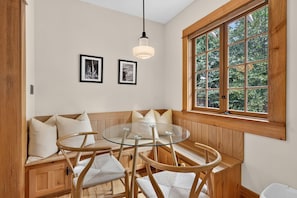 Una habitación con un banco de madera, una mesa de vidrio y sillas, una ventana con vista a árboles, y dos cuadros en la pared.