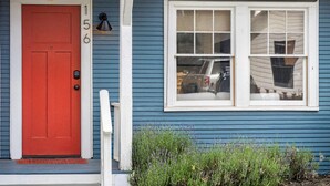 A blue house with a red door, a white window, and a small porch with a white railing.