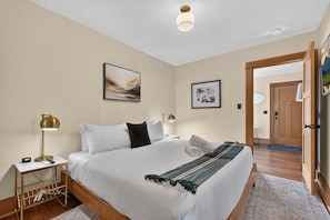 A bedroom with a bed, bedside lamps, a nightstand, a framed abstract painting, a wooden door, and a hallway.
