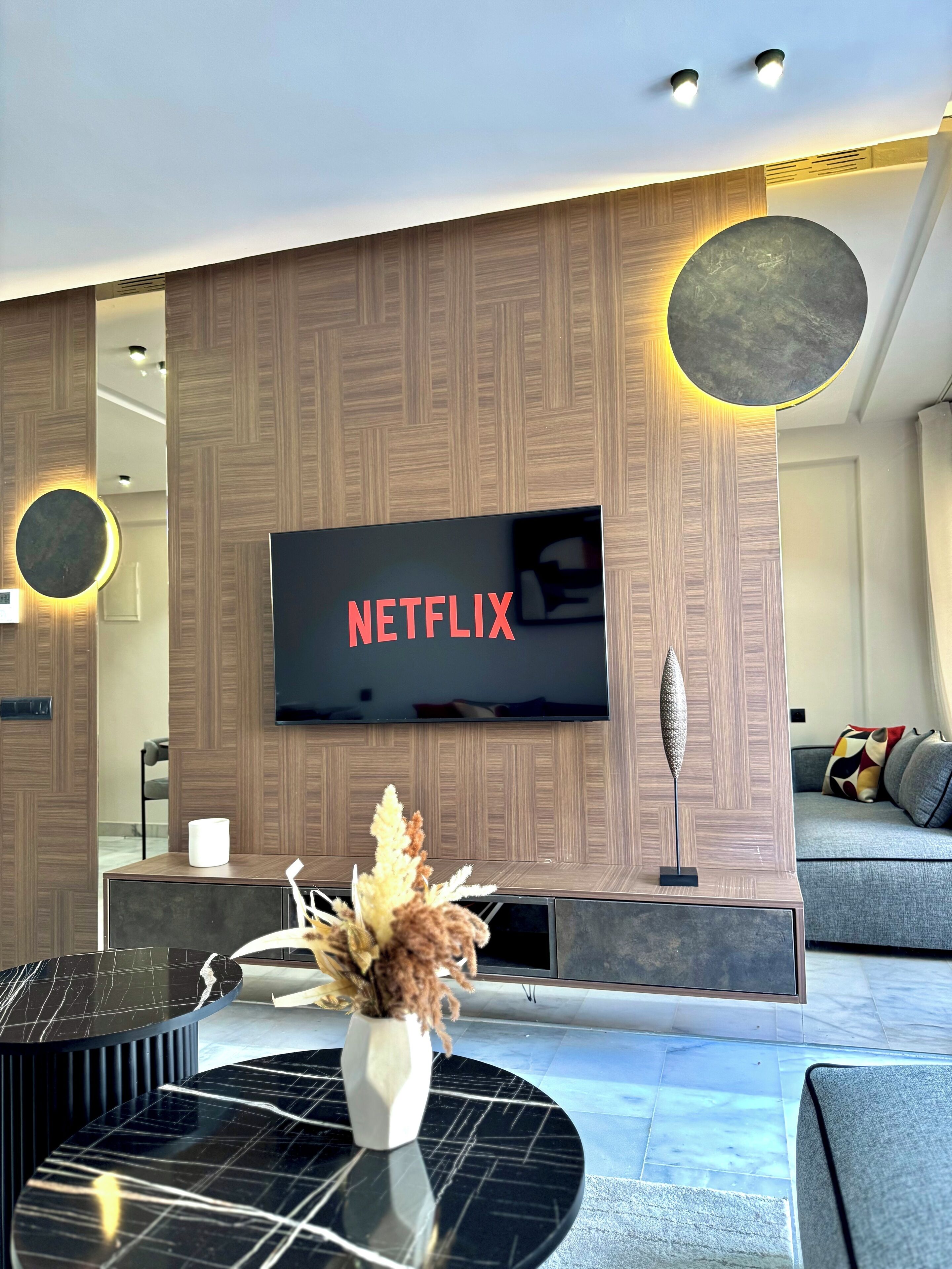 A modern living room with a flat-screen TV mounted on a wooden wall, a round table with a vase, and a sofa with cushions.
