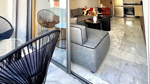 Un salon moderne avec une porte vitrée donnant sur un balcon, une cuisine équipée d’appareils électroménagers contemporains et un coin repas avec une table et des chaises en verre.
