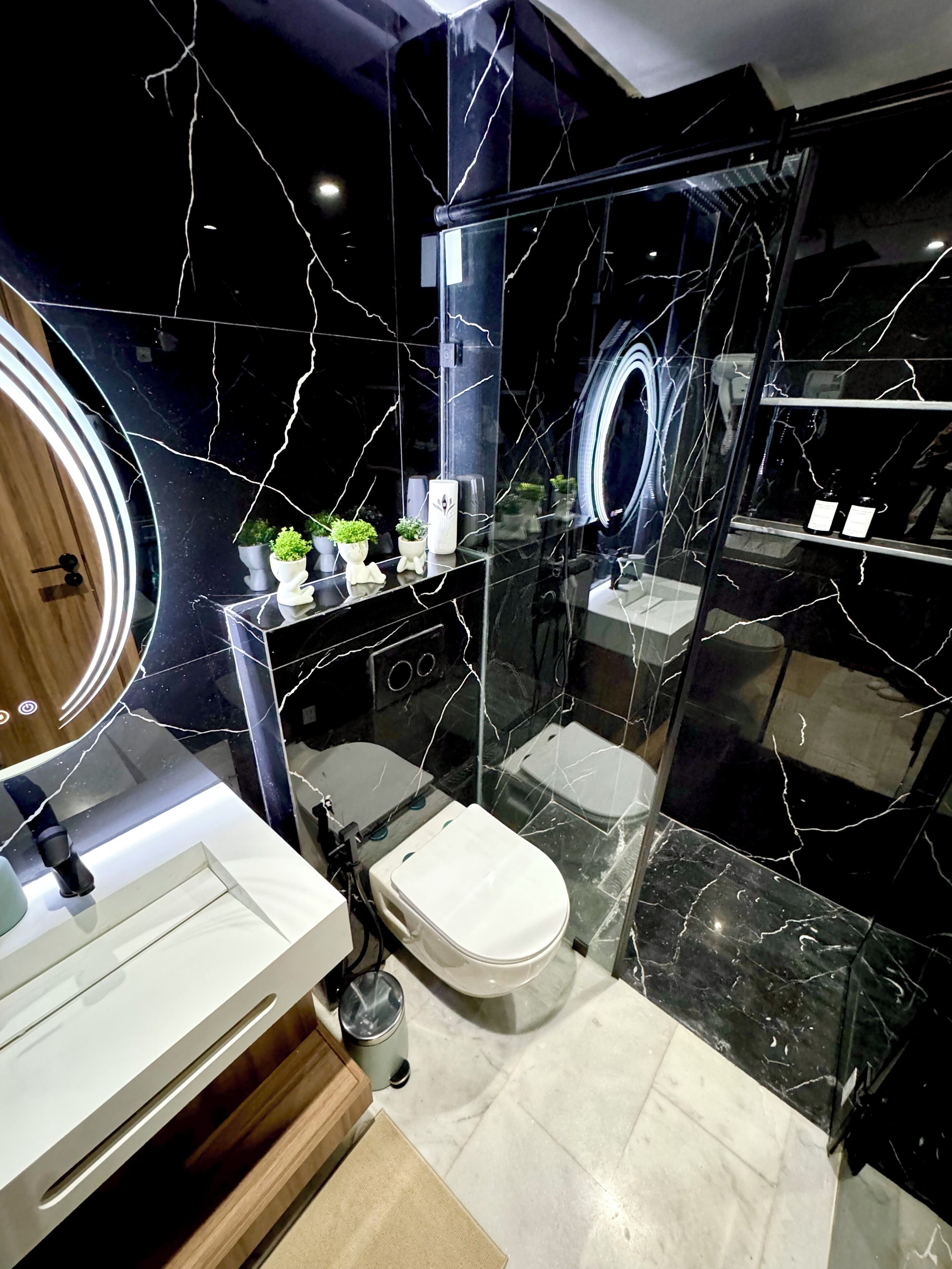 A modern bathroom with a black marble design, a glass shower enclosure, and a vanity with a mirror and plants.