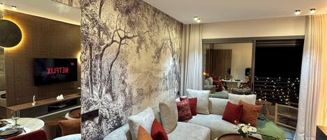 Ein modernes Wohnzimmer mit einem großen Sofa, roten Sesseln, einem Esstisch für zwei Personen und einem an der Wand montierten Fernseher.