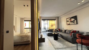 Un salon moderne avec un canapé, une table basse et un téléviseur. Une chambre adjacente contient un lit.